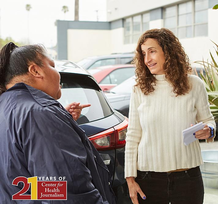 A woman with curly brown hair, wearing a white ribbed sweater, holds a notepad and pen while speaking with another person outside in a parking lot. The second person, dressed in a dark jacket, gestures with their hand while facing the woman. Cars are visible in the background, and the conversation appears informal and engaged. In the bottom-left corner, a logo reads "20 Years of Center for Health Journalism" in red and gold.