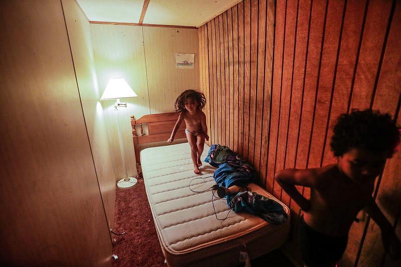 Victoria, 3, bounces on a twin bed in a room by a wall built to create an extra bedroom in her family's trailer. Her 8-year-old Romeo walks through the room.