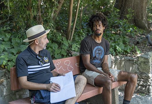 A young man sits on a park bench speaking with an older man who holds a notebook and pen. They appear to be engaged in a serious conversation surrounded by greenery.